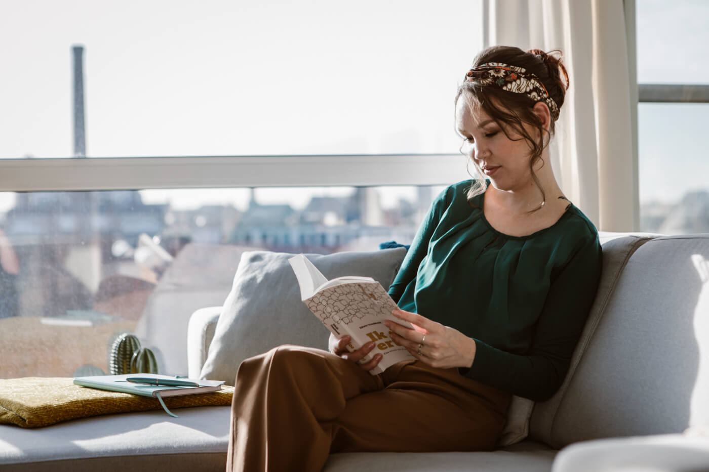 Foto door Marijn de Wijs: ik zit rechts in beeld op een lichte grijsblauwe bank. Ik ben een indo vrouw met donker opgestoken haar, ik draag een bruine broek en donkergroene blouse, in mijn haar een batik haarband. Ik lees het boek "Ik doe ertoe". Op de achtergrond is door grote ramen de stad te zien.