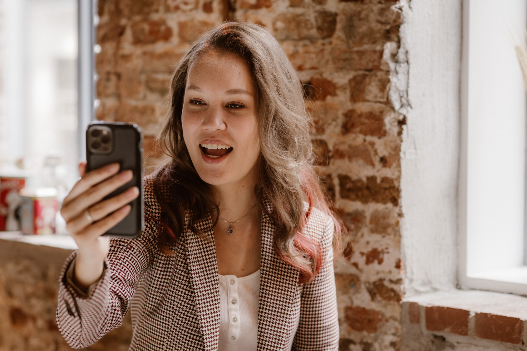 Foto door Marijn de Wijs: portret waarbij ik lachend naar een telefoon, links in beeld in mijn hand praat. Op de achtergrond een bakstenen muur met ramen. Ik ben een indo vrouw met donker golvend haar, draag een wit shirt met knoopjes, een houten kralenketting en een rood-wit-zwart geblokt colbert. 