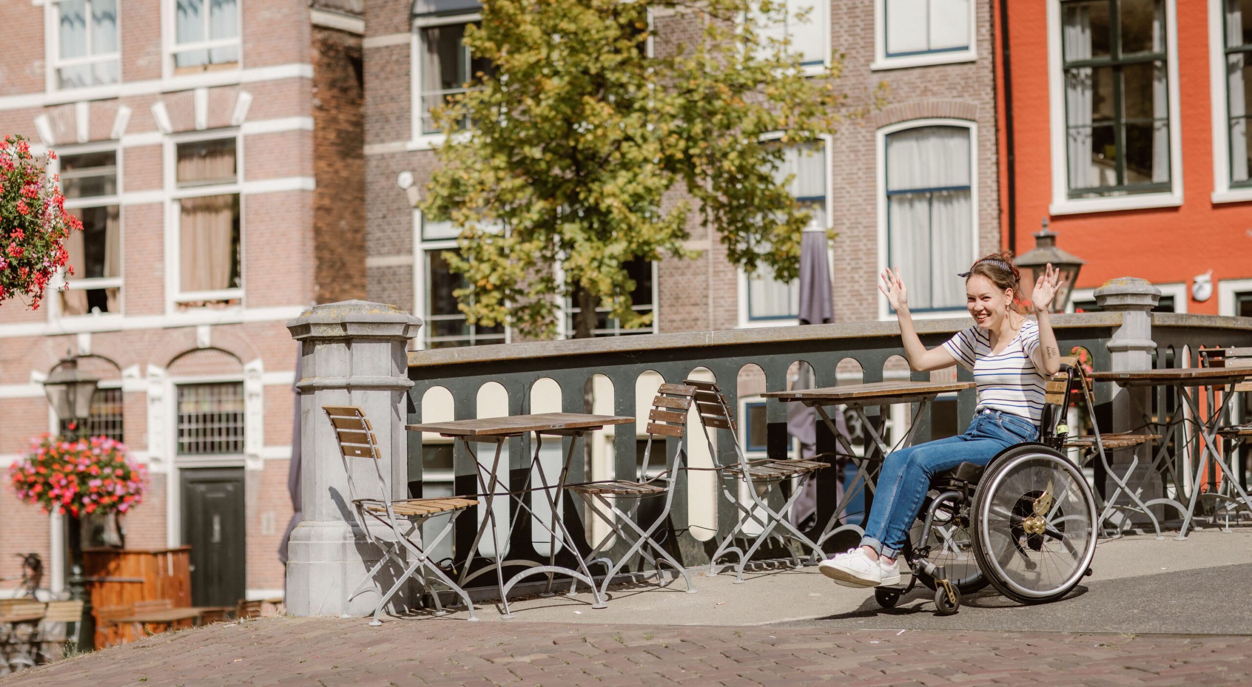 Foto van Marijn de Wijs; een stuk brug in Leiden. Rechts in beeld kom ik van de brug af gerold in mijn rolstoel. Ik ben een indo vrouw met opgestoken donker haar. Ik draag een hoge spijkerbroek en wit met blauw-gestreept shirt. Ik lach naar de camera en houd mijn handen in de lucht. Op de achtergrond zijn panden te zien en op de brug staan een paar terrastafels met stoelen.
