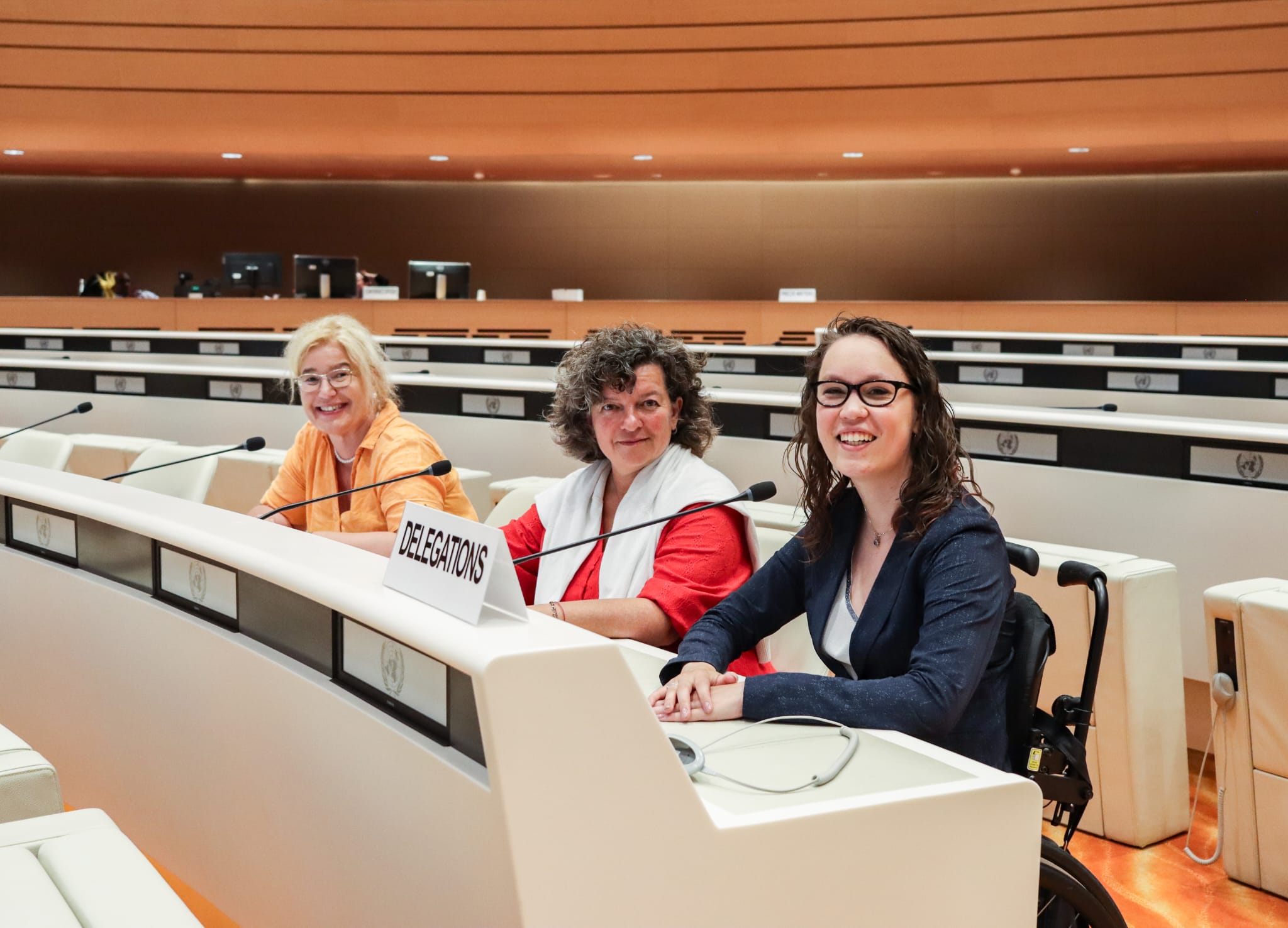 Foto door Jens Van Hecke: Roos Hoelen, Illya Soffer en ik achter de crèmewitte desk van een VN-zaal. Roos is een witte vrouw met blond haar, een dun montuur bril en een oranje jurk. Illya is een witte vrouw met donker en grijs krullend haar, een rode jurk en daarover losjes een wit vest over haar schouders. Ik ben een indo vrouw met donker golvend haar en een bruin montuur bril op. Ik draag een witte blouse met glinsterend zilveren bies en een donkerblauw colbert. We kijken allen lachend schuin naar de camera en hebben ieder een kleine microfoon voor ons.