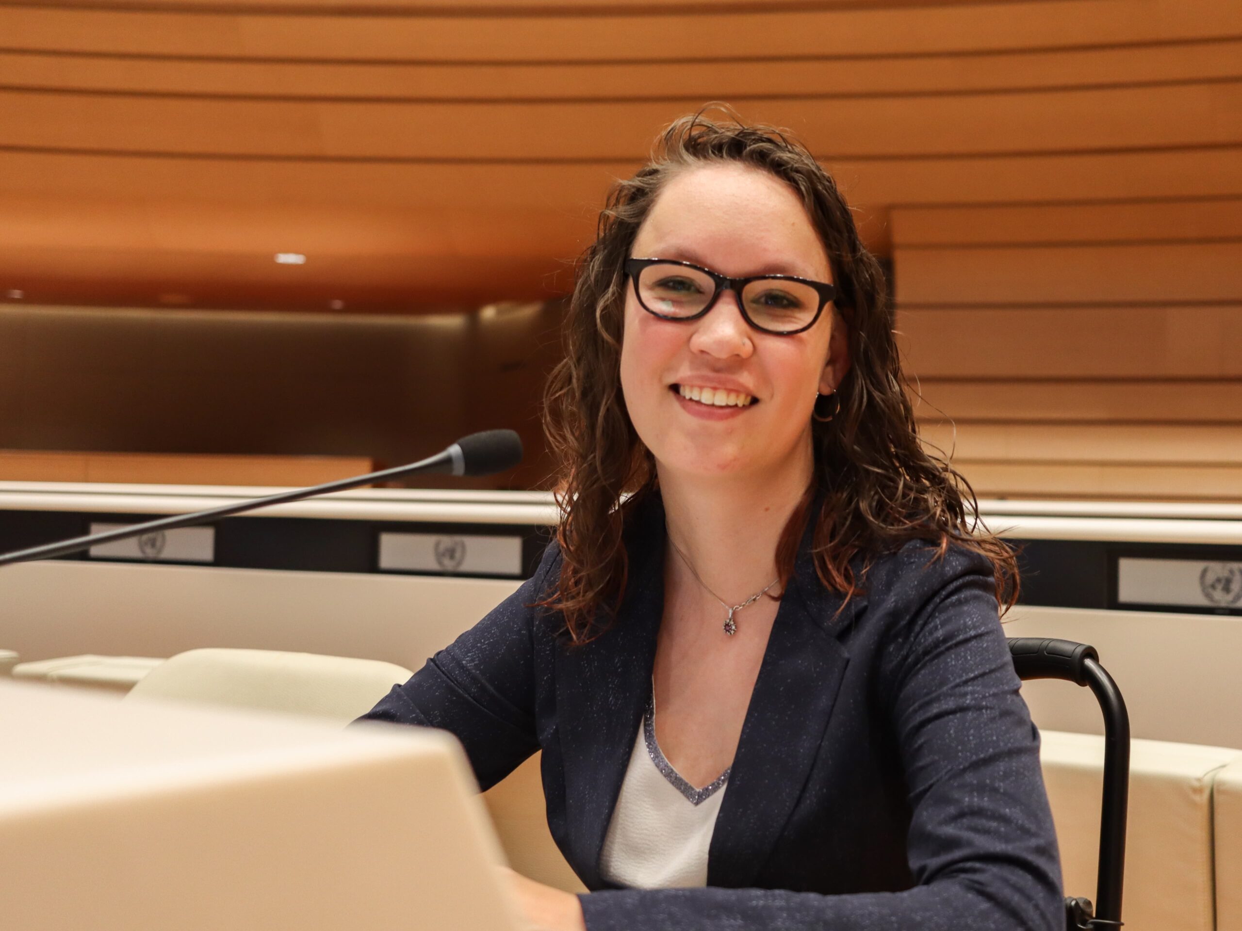 Foto door Jens Van Hecke: ik zit in mijn rolstoel achter de crèmewitte desk van een VN-zaal. Ik ben een indo vrouw met donker golvend haar en een bruin montuur bril op. Ik draag een witte blouse met glinsterend zilveren bies en een donkerblauw colbert. Ik kijk schuin lachend naar de camera, links van mij is een microfoon te zien. 