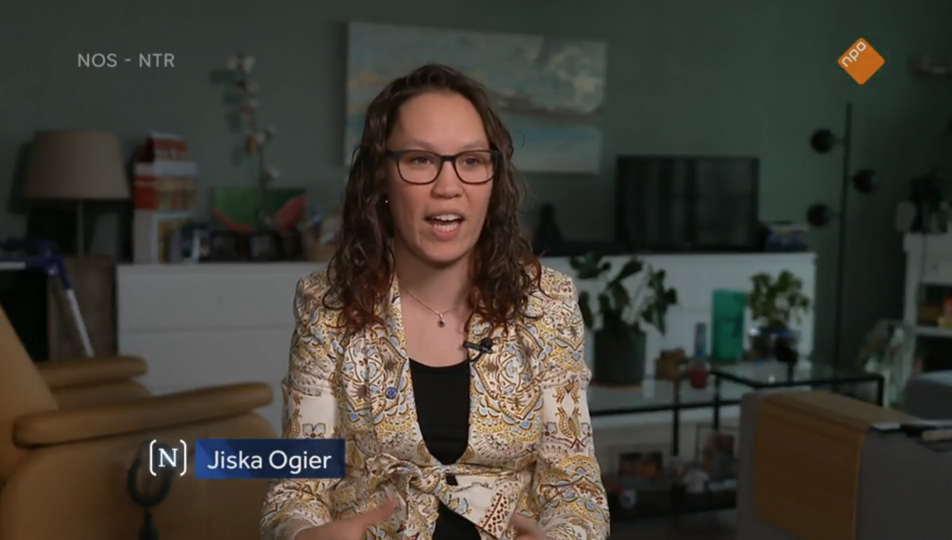 Beeld uit de uitzending van Nieuwsuur. Ik zit op de voorgrond in mijn woning, ik ben een indo vrouw met donker golvend haar en een donker montuur bril. Ik draag een zwart shirt met een vrolijk meerkleurig colbert met op het revers een speldje van de VN. Links onderin beeld het logo van Nieuwsuur en de balk met mijn naam Jiska Ogier. Linksboven NOS-NTR en rechtsboven het logo van NPO (oranje). 