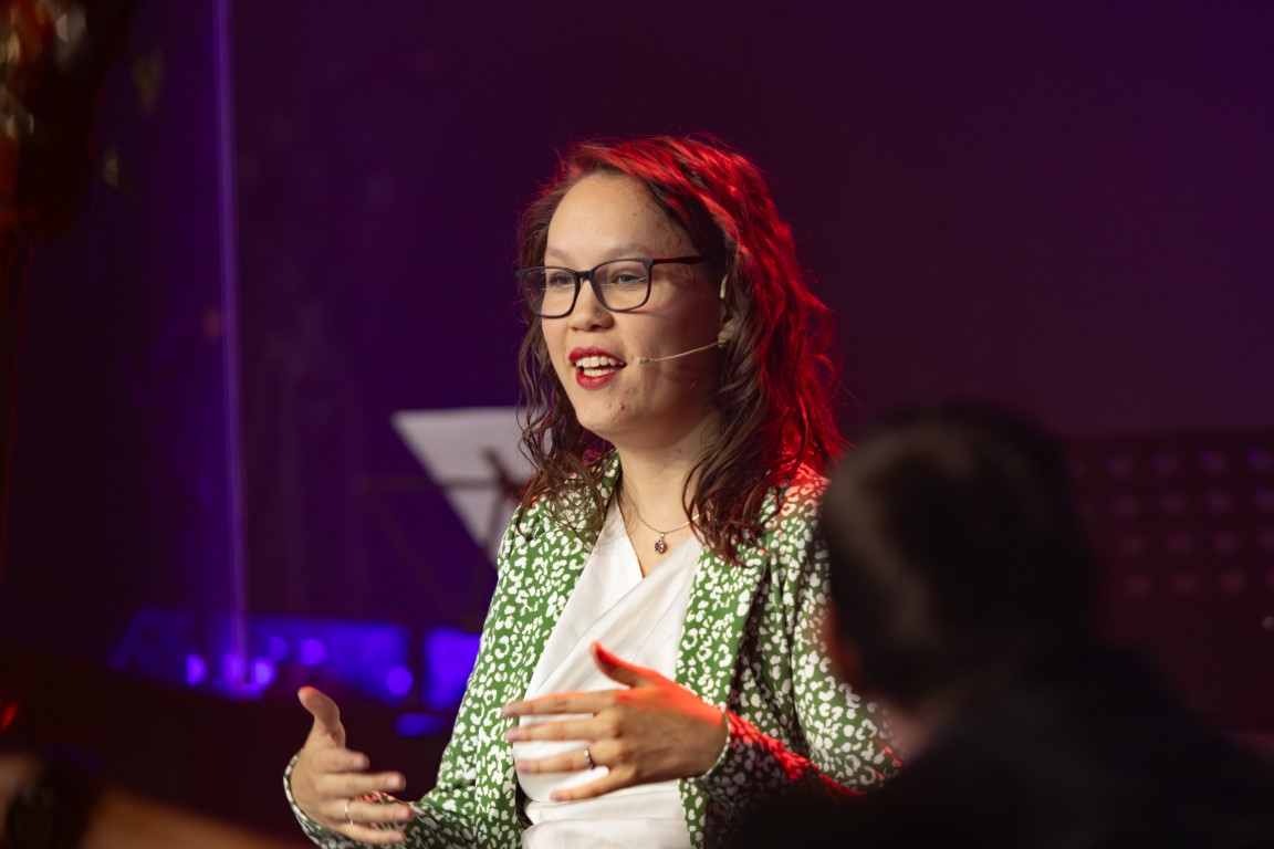 Foto door Jens van Hecke; ik zit rechts in beeld en praat naar links in een microfoon. Ik ben een indo vrouw met opgestoken donker haar met een batik haarband erin. Ik draag een oranje sjaal en donkerblauwe jas. 