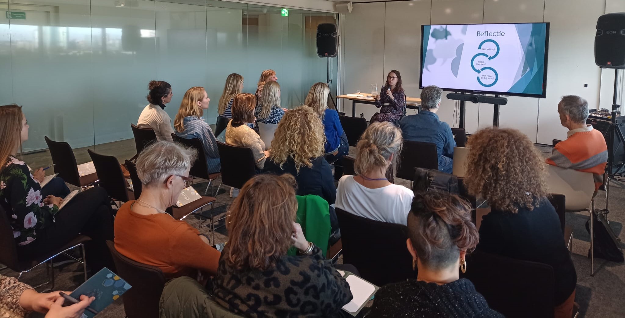 Foto door Jeroen Havers, schuin achterin de zaal genomen. Je ziet de ruggen en zijkanten van publiek, vooraan de zaal sta ik. Ik ben een indo vrouw met donker golvend haar en een donker montuur bril, ik zit in een zwarte rolstoel en houd een microfoon vast. 