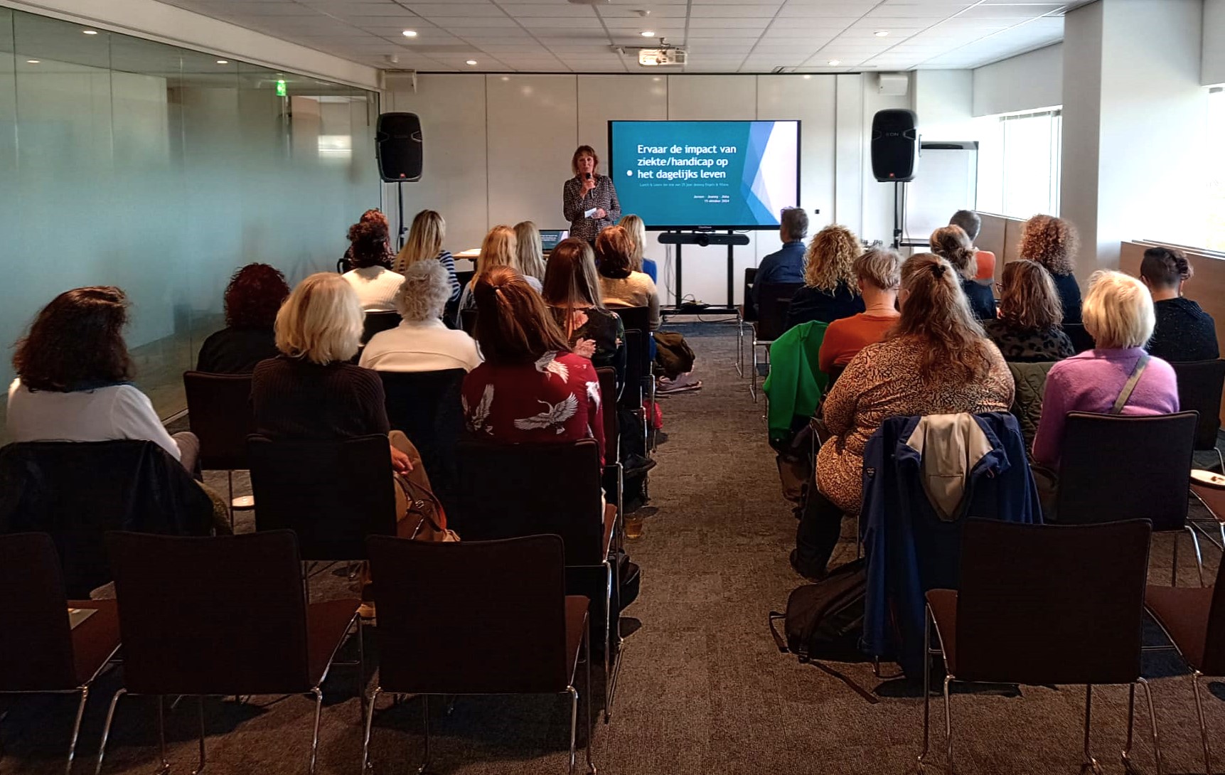 Foto door Jeroen Havers, genomen achterin de zaal. In beeld zijn de ruggen van het publiek, vooraan staat Jeanny Engels voor de presentatie. Jeanny is een witte vrouw met bruin haar en draagt een blouse met motief en houd de microfoon in haar rechter hand. In haar linker hand heeft ze een klein briefje.