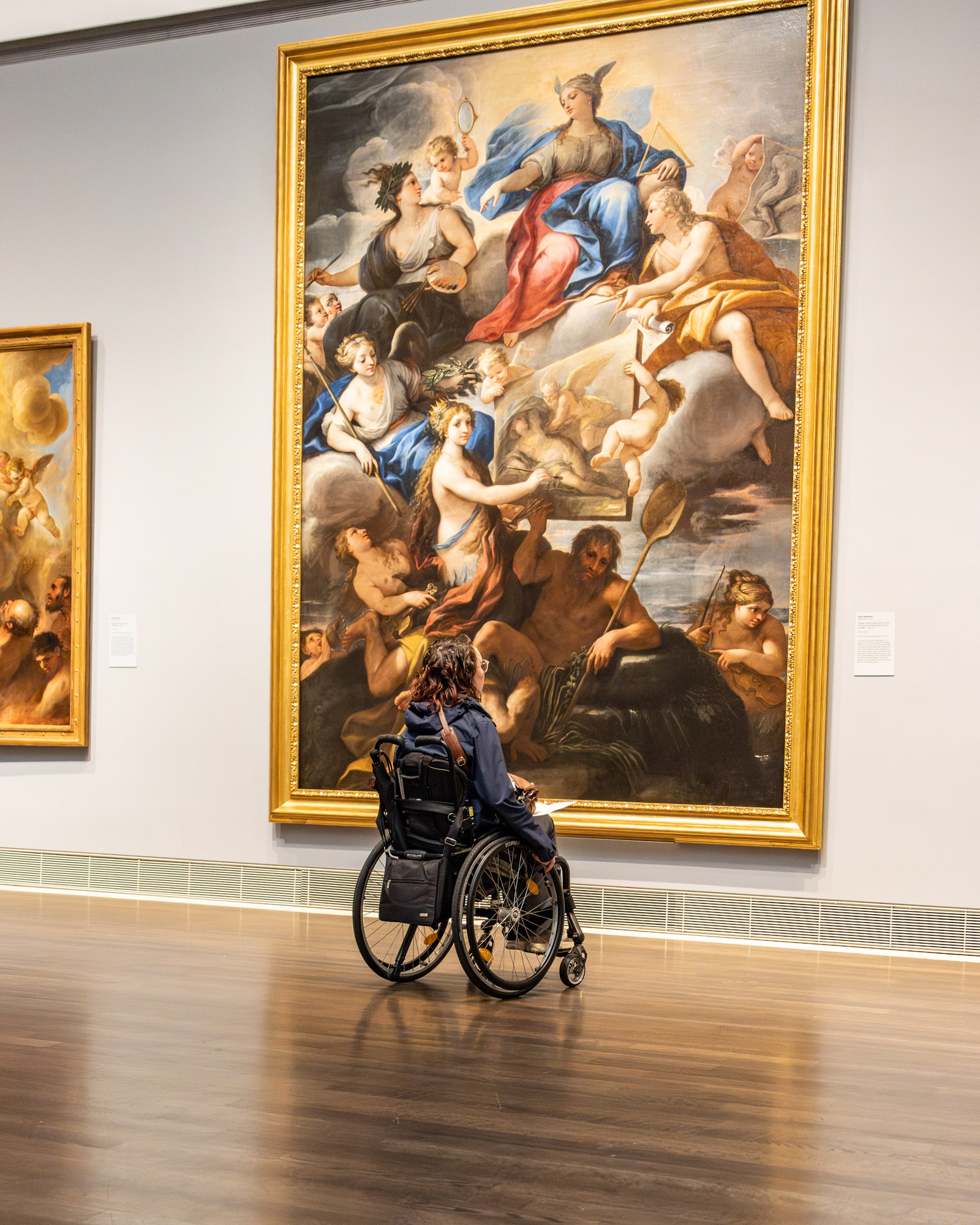 Foto door Jens Van Hecke: ik sta met mijn rug naar de camera naar een enorm schilderij te kijken. Ik ben een indo vrouw met donker golvend haar tot schouderlengte en zit in een zwarte rolstoel. Ik sta op een glimmende houten vloer en kijk naar een metershoog schilderij met engelachtige figuren aan de muur. 