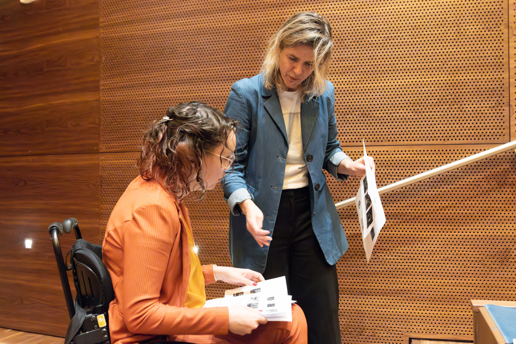 Foto door Jens Van Hecke: ik sta op de voorgrond in mijn zwarte rolstoel, achter mij staat Cathelijne. Ik ben een indo vrouw met donker golvend haar en een transparant montuur bril. Ik draag een okergele blouse en een oranje pak. Op mijn schoot ligt papier waar ik naar kijk. Cathelijne is een witte vrouw met blond haar, ze draagt een donkere broek, wit T-shirt en denimblauw lang colbert. In haar ene hand heeft ze een stapel papier, met de andere wijst ze naar de papiere op mijn schoot. We zijn in gesprek. 