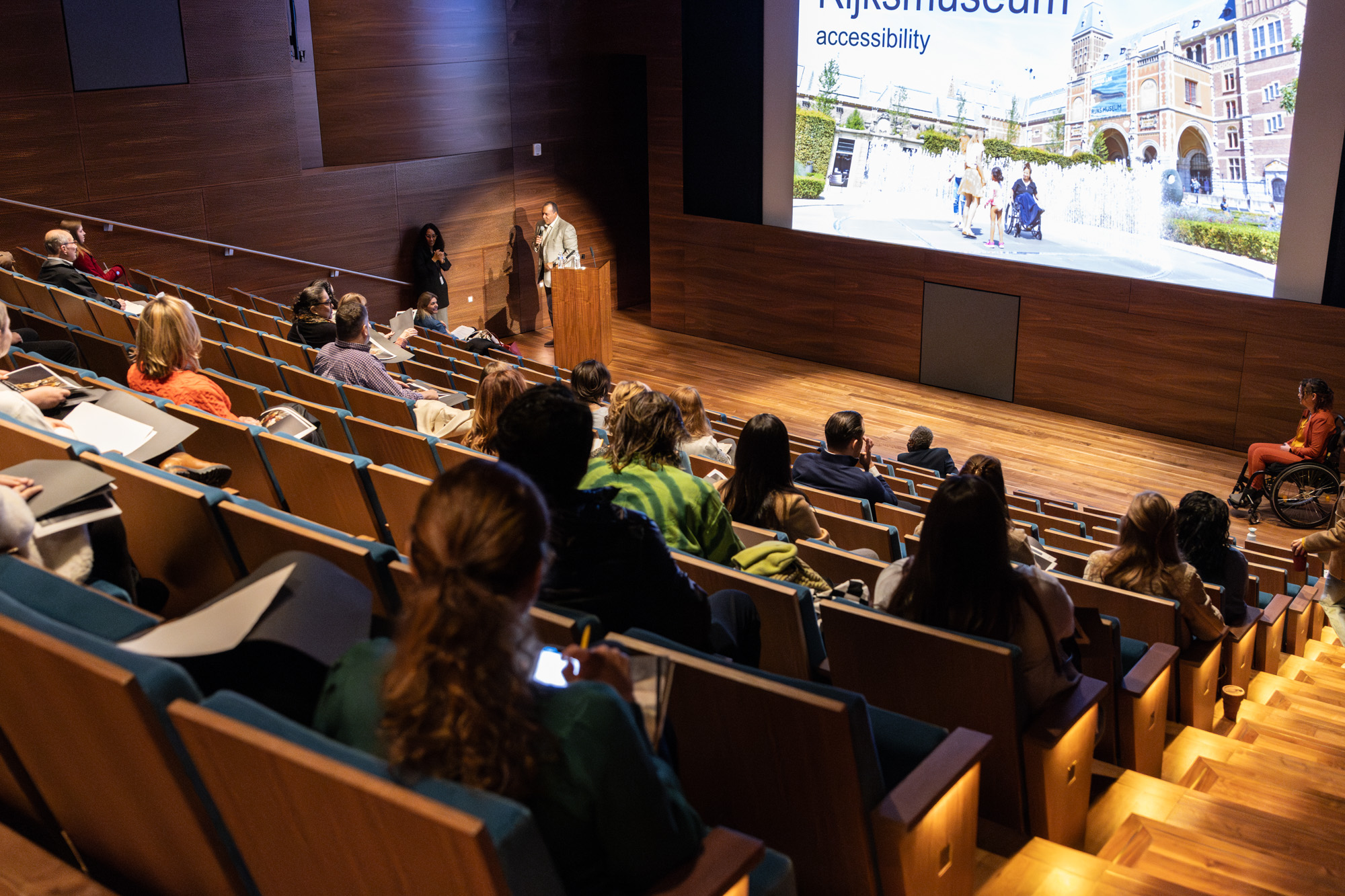 Foto door Jens Van Hecke: zaal in theateropstelling van bovenaf gefotografeert, in de stoelen zitten mensen. Voor aan de zaal op een groot scherm de PowerPointpresentatie. Links in beeld loopt Larry van het spreekgestoelte weg (donkere man, wit overhemd, grijs colbert), rechts in beeld zit ik voor het scherm (indo vrouw, zwarte rolstoel, oranje pak). 