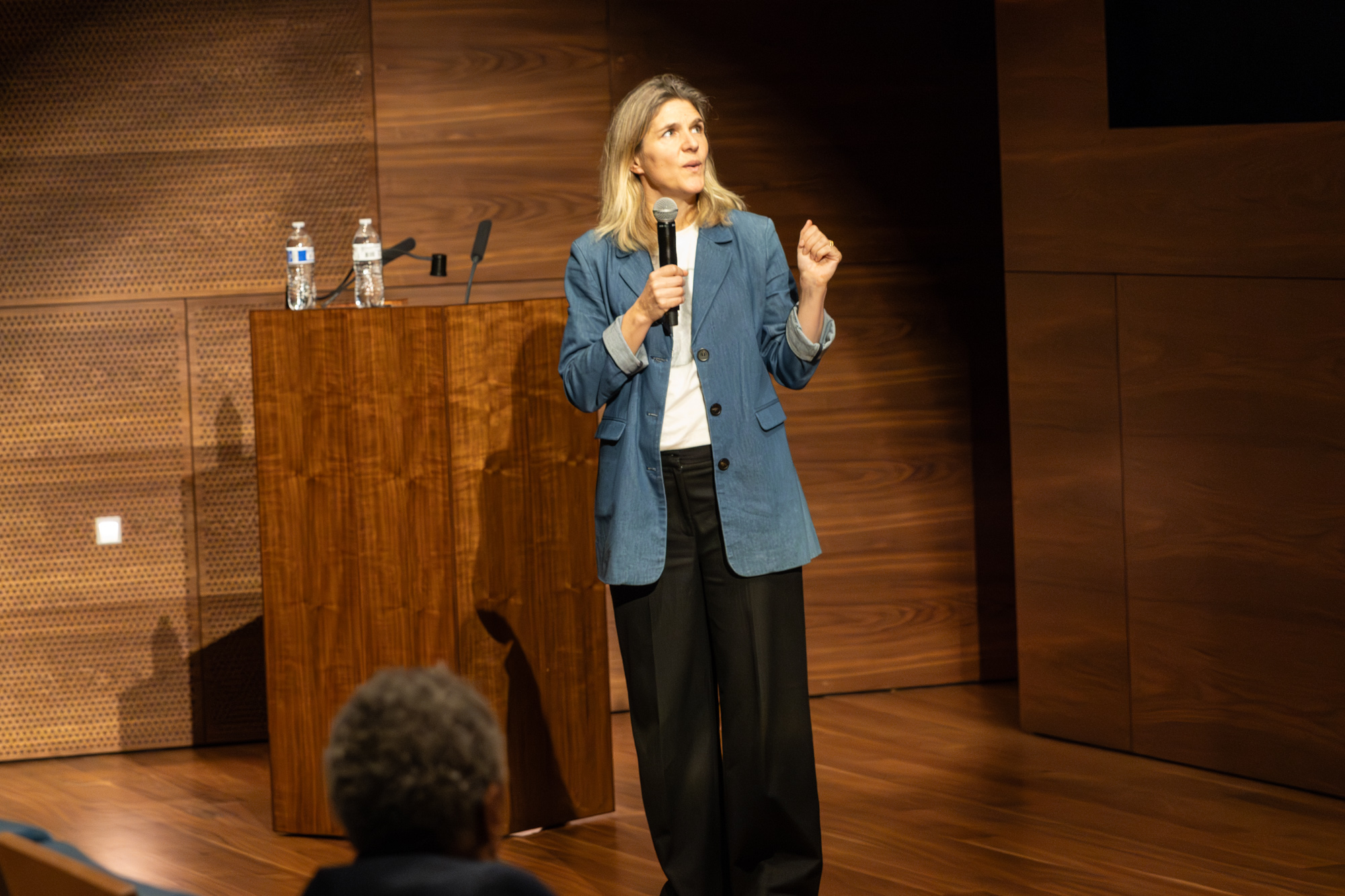 Foto door Jens Van Hecke: in een donkere zaal staat Cathelijne met een microfoon in haar hand te praten. Cathelijne is een witte vrouw met blond haar, ze draagt een donkere broek met een wit T-shirt en een denimblauw lang colbert. Ze kijkt naar rechts buiten beeld en gebaart met een hand. Op de voorgrond is het achterhoofd van een toehoorder zichtbaar. 