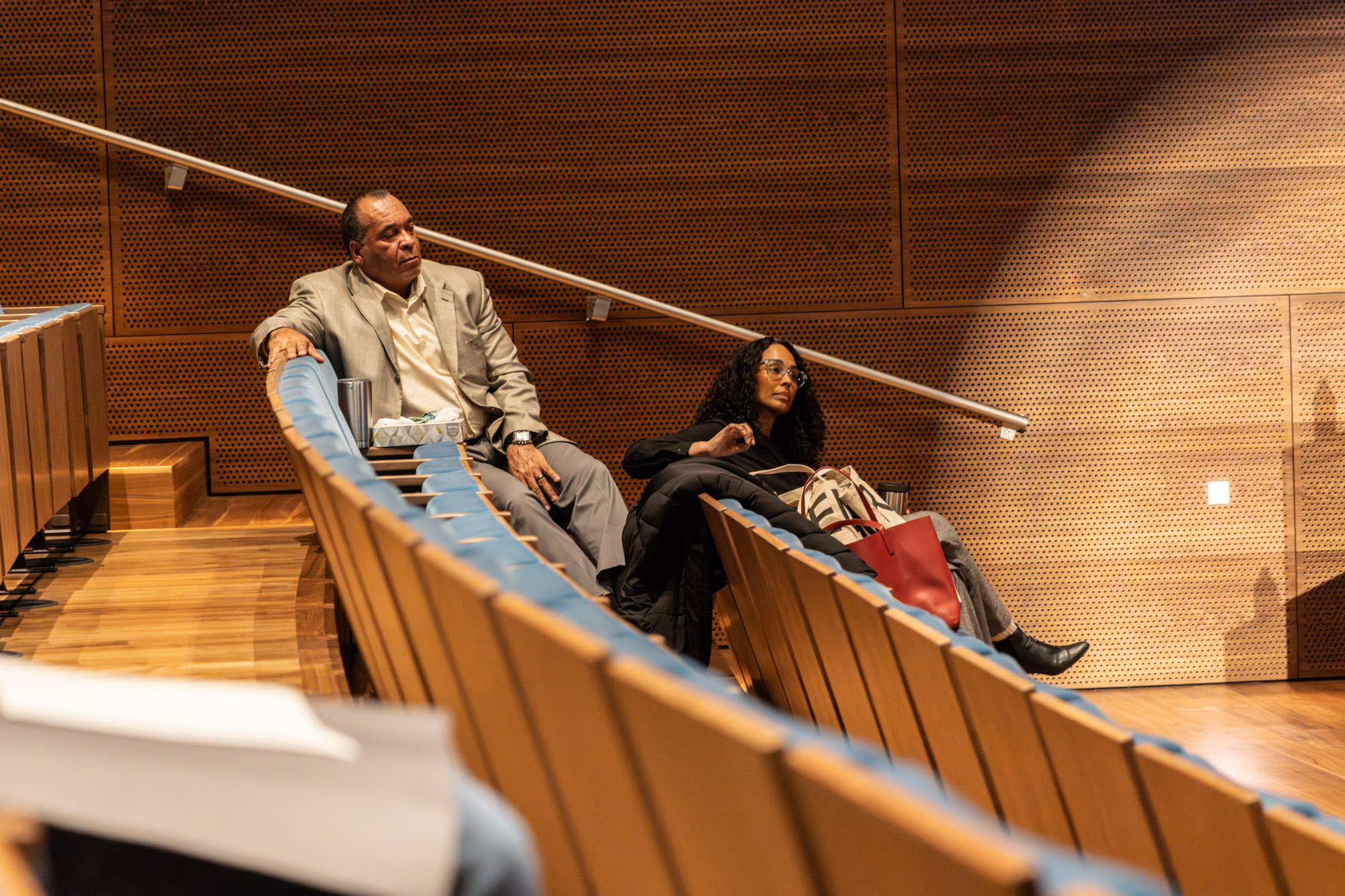 Foto door Jens Van Hecke: deel van de theaterzaal met achterin twee toehoorders. Larry, een donkere man die een wit overhemd en grijs colbert draagt. En Leeza, een donkere vrouw met donkere krullen en een transparant groot monuur bril. Ze kijken naar rechts buiten beeld, waar de sprekers staan. 