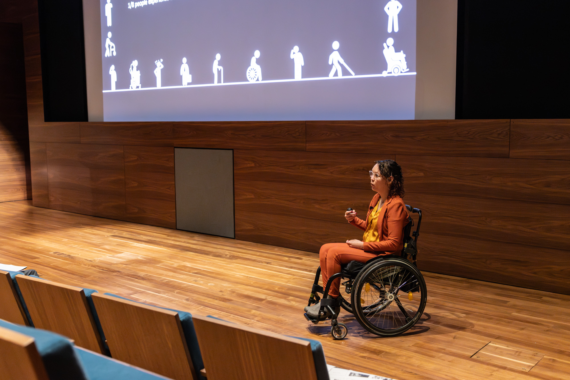 Foto door Jens Van Hecke: donkere zaal met op de voorgrond links nog een aantal stoelen zichtbaar. Ik sta rechts in beeld; indo vrouw met donker golvend haar, ik draag een glimmende okergele blouse en oranje pak, ik zit in een zwarte rolstoel en gebaar met mijn handen. Op de achtergrond boven mij is een stukje van de PowerPoint zichtbaar, een donker vlak met daarop witte figuurtjes met allerlei soorten (on)zichtbare handicaps. 