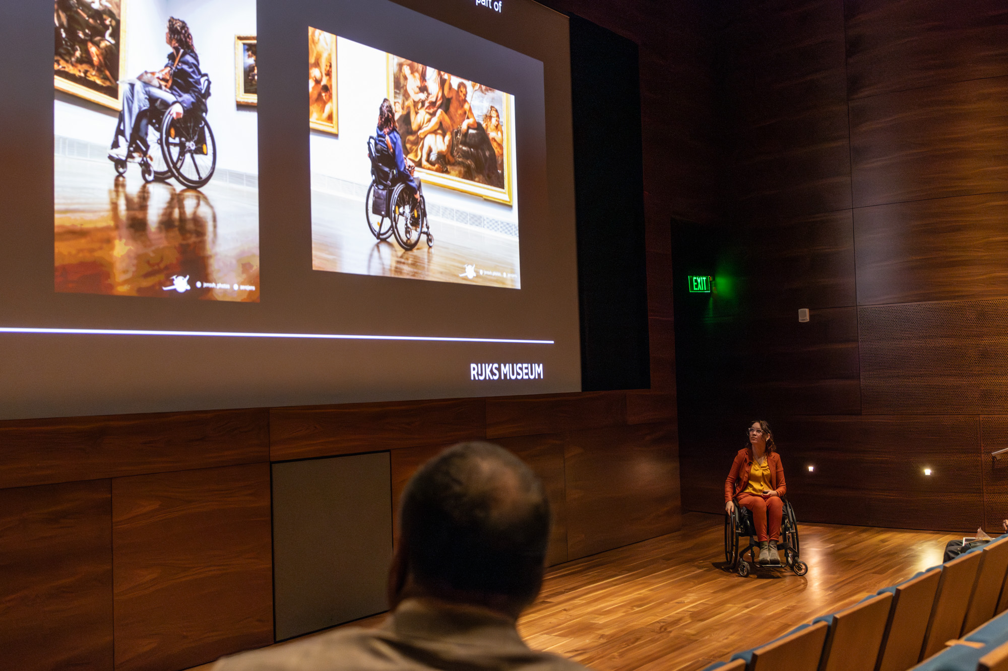 Foto door Jens Van Hecke: de donkerbruine zaal waarin de trainingen plaatsvonden. Links op de voorgrond het achterhoofd van een toehoorder. Links groot op het scherm een donkere presentatie met daarop twee foto's van mijzelf in het Museum of Fine Arts Houston, vanaf mijn rug gefotografeerd terwijl ik naar schilderijen kijk. Rechts onder het scherm sta ik; indo vrouw met donker golvend haar en transparant montuur bril, glimmende okergele blouse en oranje pak, in mijn zwarte rolstoel. 