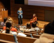 Foto door Jens Van Hecke: donkere zaal met op de voorgrond een theateropstelling van stoelen waar deels mensen in zitten. Beneden op het podium staan Cathelijne en Jiska. Cathelijne is een witte vrouw met blond haar, ze draagt een donkere broek, een wit T-shirt en een denim-blauw lang colbert, ze kijkt naar Jiska. Jiska is een indo vrouw met donker golvend schouderlengte haar in een zwarte rolstoel. Ze draagt een okergele top met een oranje pak en een transparante bril.