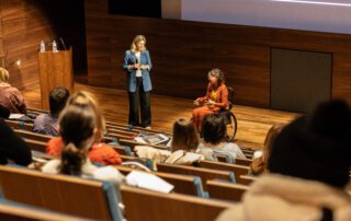 Foto door Jens Van Hecke: donkere zaal met op de voorgrond een theateropstelling van stoelen waar deels mensen in zitten. Beneden op het podium staan Cathelijne en Jiska. Cathelijne is een witte vrouw met blond haar, ze draagt een donkere broek, een wit T-shirt en een denim-blauw lang colbert, ze kijkt naar Jiska. Jiska is een indo vrouw met donker golvend schouderlengte haar in een zwarte rolstoel. Ze draagt een okergele top met een oranje pak en een transparante bril.