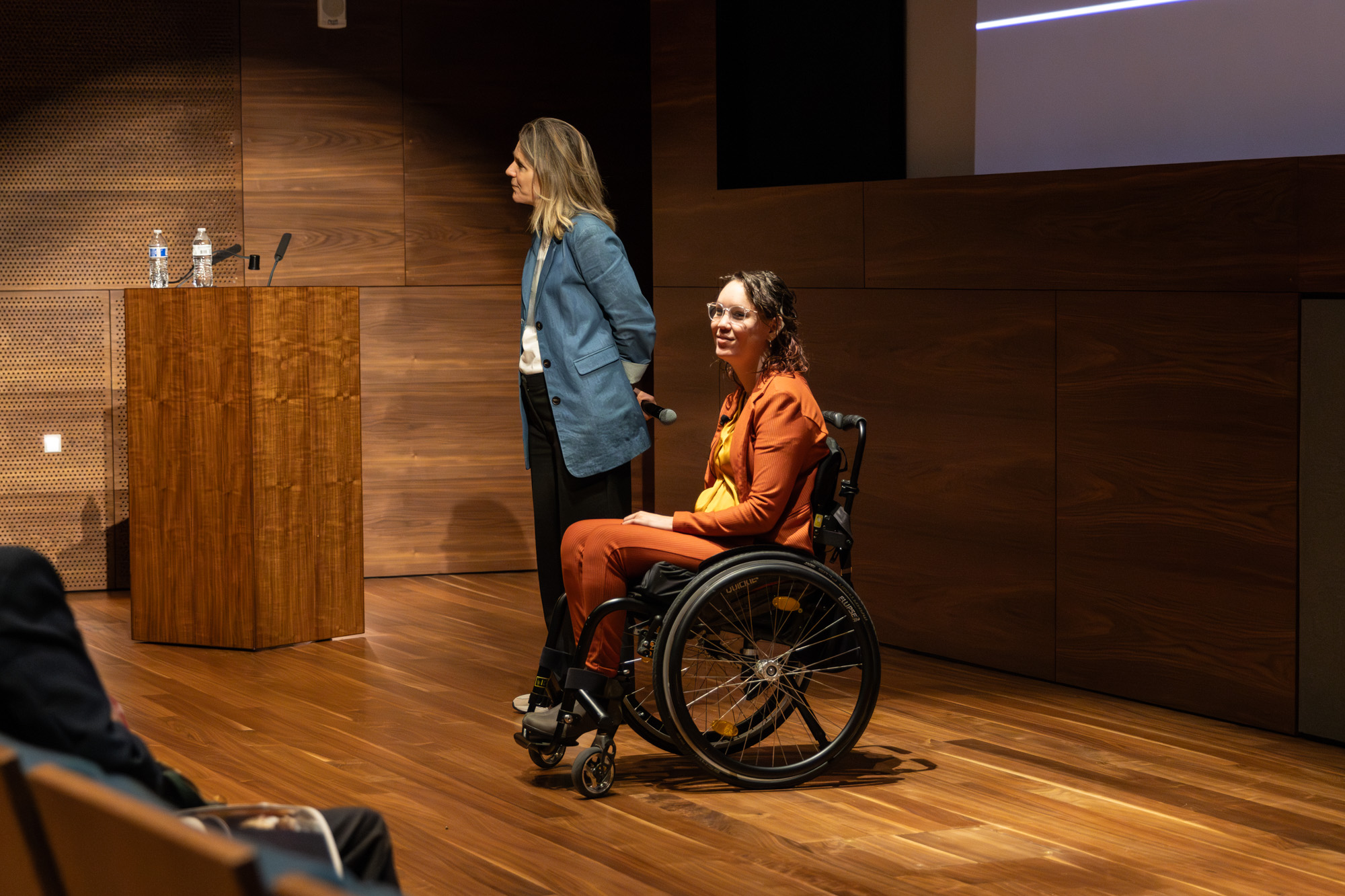 Foto door Jens Van Hecke: donkere zaal met links in beeld stoelen en toehoorders. Midden in beeld staan Cathelijne en ik, we kijken naar links (de zaal) en luisteren naar een reactie uit de zaal. Cathelijne is een witte vrouw met blond haar, ze draagt een donkere broek, wit T-shirt en denimblauw lang colbert en heeft haar handen op haar rug. Ik ben een indo vrouw met donker golvend haar, draag een glimmende okergele blouse en oranje pak en zit in een zwarte rolstoel. 