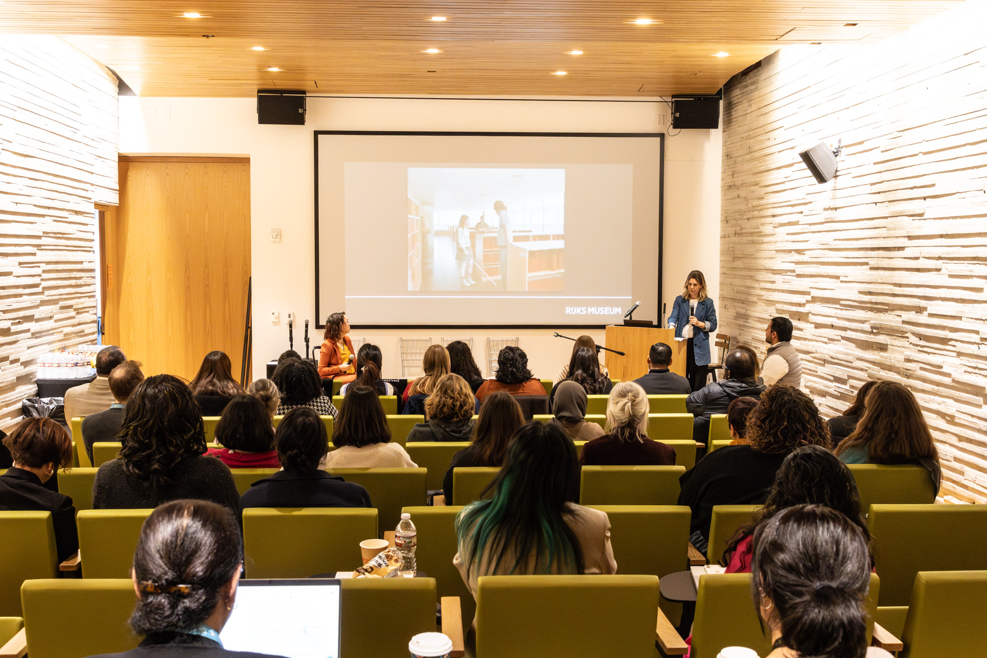 Foto door Jens Van Hecke: symposiumzaal van achterin gefotografeerd. Op de voorgrond mensen in stoelen, op de achtergrond van de foto een groot scherm met een PowerPointpresentatie, links in beeld ben ik te zien. Een indo vrouw met donker golvend haar in een zwarte rolstoel met een oranje pak. Rechts in beeld Cathelijne, een witte vrouw met blond haar, een donkere broek, wit T-shirt en een denimblauw lang colbert. 