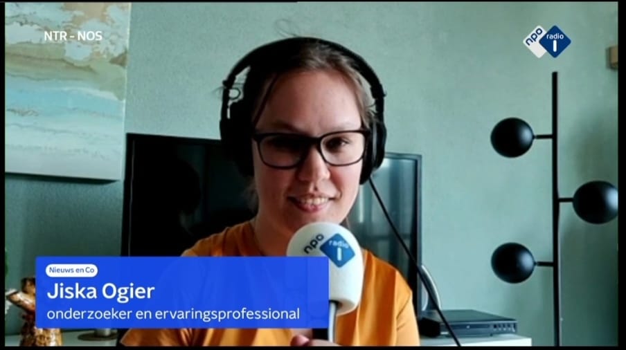 Stil uit het videobeeld van de uitzending van Nieuws & Co. Ik ben een indo vrouw met donker haar in een staartje, ik draag een bruin montuur bril, koptelefoon en okergele blouse. Voor mijn gezicht heb ik een microfoon met NPO Radio 1 erop. Links in beeld een blauwe balk van Nieuws & Co met de tekst "Jiska Ogier, onderzoeker en ervaringsprofessional".