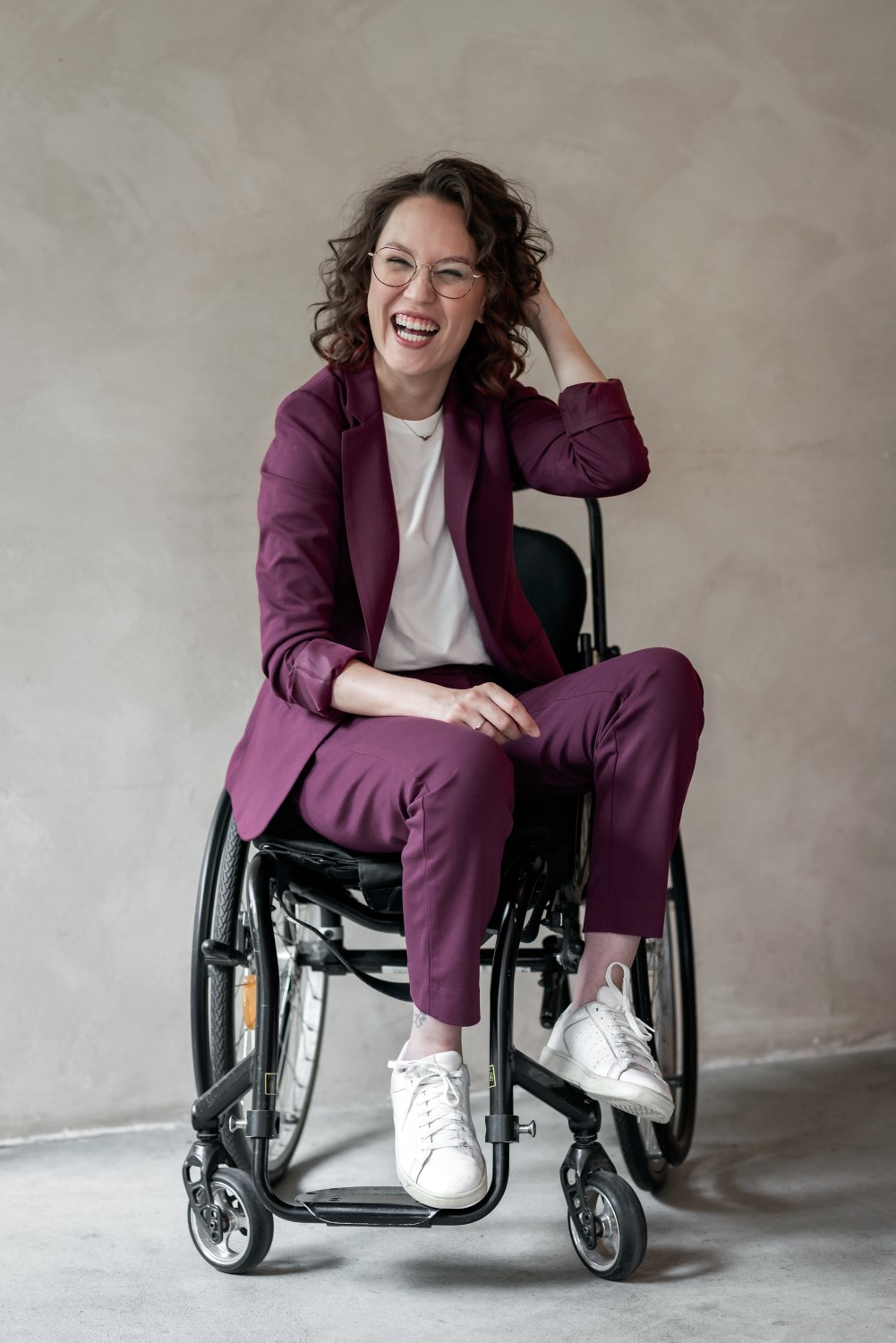 Foto door Marijn de Wijs van mij. Ik ben een indo vrouw met donker golvend haar. Ik zit schuin in een zwarte rolstoel, draag een bordeaux rood broekpak met witte sneakers en een wit t-shirt. Ik lach uitbundig met één hand in mijn haar. 