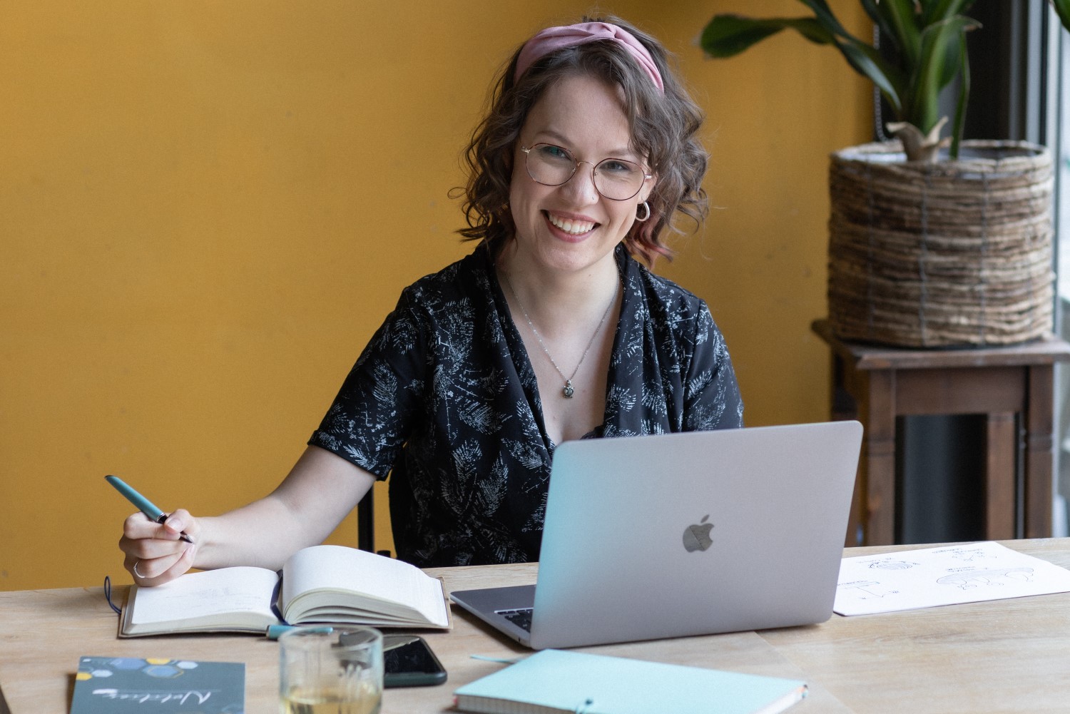 Foto door Marijn de Wijs: ik zit aan een bureau met schrijfboek en laptop. Ik ben een indo vrouw met donker golvend haar en lichtbruin montuur bril. Ik draag een zwart-witte jumpsuit en roze haarband. Ik kijk op naar de camera met een lach, in mijn hand heb ik een pen die boven het papier zweeft.
