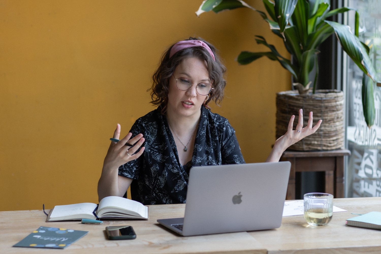 Foto door Marijn de Wijs: Ik zit aan een bureau met een schrijfboek en laptop. Ik ben een indo vrouw met donker golvend haar en lichtbruin montuur bril, ik draag een zwart-witte jumpsuit en roze haarband. Ik praat naar het scherm en beweeg met mijn handen.