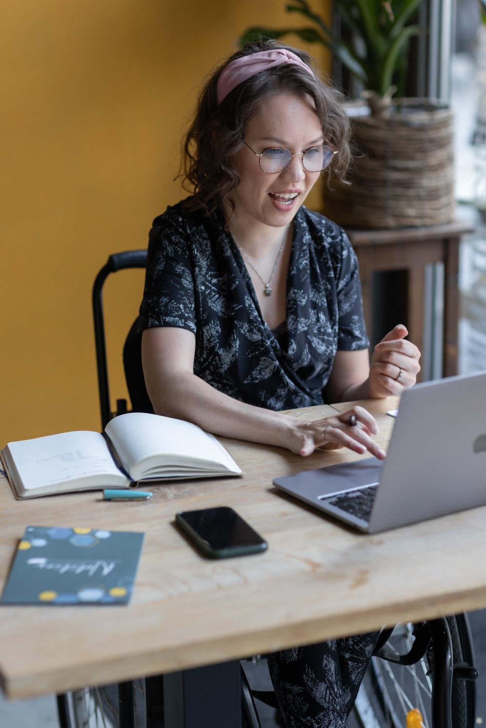 digi-MarijndeWijs_25-115 Foto door Marijn de Wijs: ik ben een indo vrouw met donker golvend haar en een roze haarband, ik draag een dun montuur bruine bril en een zwart-witte jumpsuit. Achter mij een okergele muur en planten. Ik zit in mijn rolstoel aan een bureau, praat naar mijn laptop met mijn hand naar het toetsenbord. Naast mij ligt een opengeslagen schrijfboek, een folder in mijn huisstijl en een telefoon.