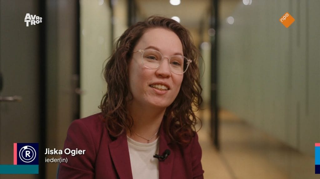 Stil beeld uit de Radar uitzending. Portret van mij in beeld, ik ben een indo met donker golvend haar en een bril, ik draag een donkerrood pak met wit shirt en praat richting de camera. Links onderin beeld het logo van Radar, "Jiska Ogier" en "Ieder(in)"