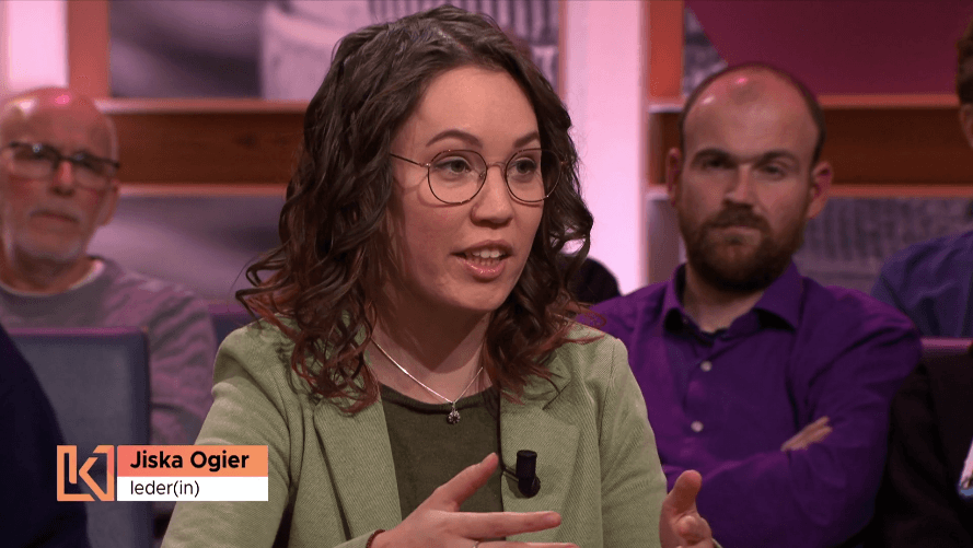 Still uit de uitzending van Kassa. Ik ben een indo vrouw met donker golvend haar en draag een dun bruin montuur bril. Ik heb een lichtgroen pak met donkergroene trui aan, ik praat richting de presentatrice. Achter in beeld zit publiek. Links een naambalkje met "Jiska Ogier" en "Ieder(in)" met het logo van Kassa.