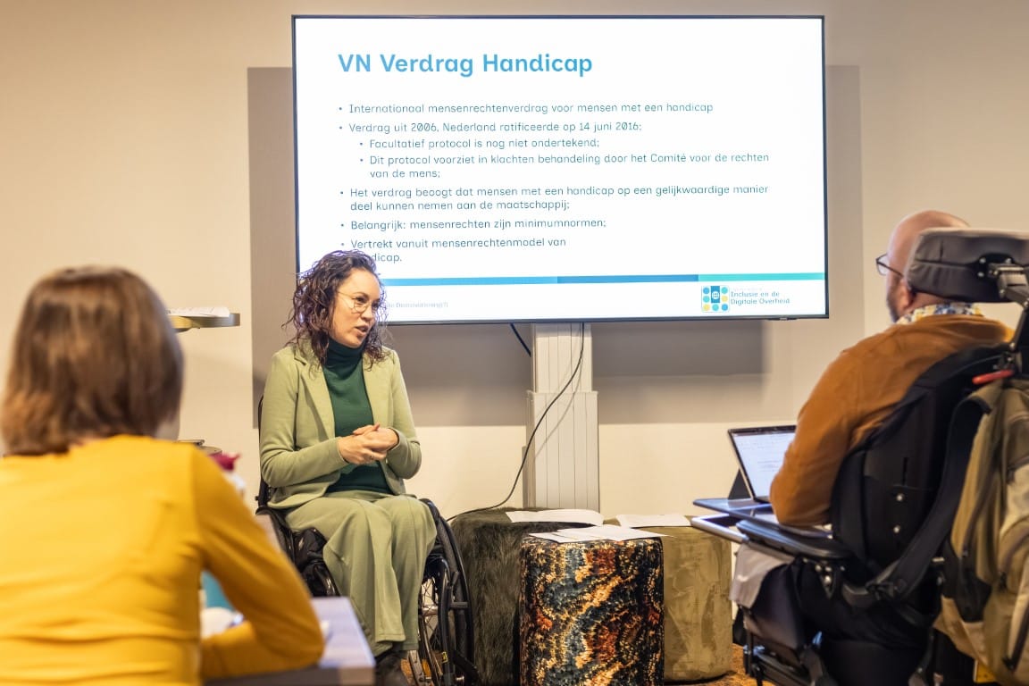Jiska is te zien tijdens de presentatie. Op de voorgrond staan deelnemers, op de achtergrond een groot scherm met de powerpoint. Daarop staat informatie over het VN-verdrag Handicap. Zij is een indo vrouw met donker golvend haar en zit in een rolstoel. 
