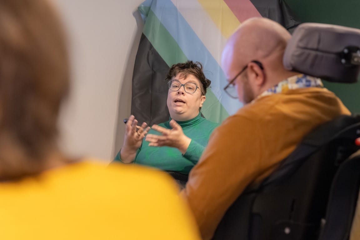 Foto van de lancering, Maranke is tussen deelnemers door te zien. Op de achtergrond hangt een disability pride flag. Die is een wit persoon zittend in een rolstoel en praat met diens handen iets uitbeeldend.