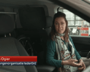Still uit NOS journaal reportage. Ik ben een indo vrouw met donker golvend haar en zit in mijn auto achter het stuur. Ik draag een dun montuur bril, donker turqoise blouse en mintgroene batik sjaal. Links in beeld een rode en blauwe balk met "Jiska Ogier" en "Belangenbehartiger Ieder(in)".