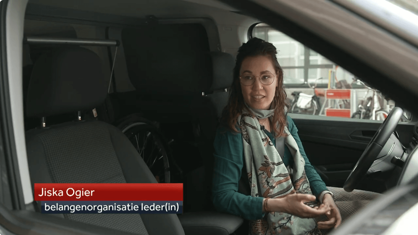 Still uit NOS journaal reportage. Ik ben een indo vrouw met donker golvend haar en zit in mijn auto achter het stuur. Ik draag een dun montuur bril, donker turqoise blouse en mintgroene batik sjaal. Links in beeld een rode en blauwe balk met "Jiska Ogier" en "Belangenbehartiger Ieder(in)".